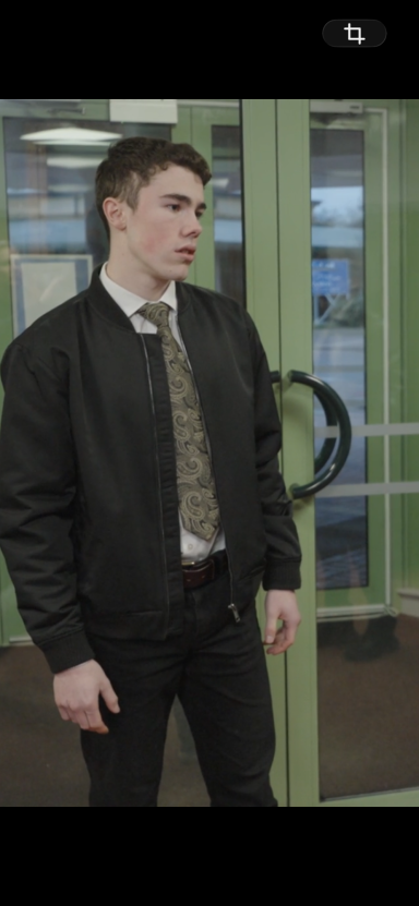 Robby McKay Young man in a black bomber jacket and tie, standing in an indoor setting.