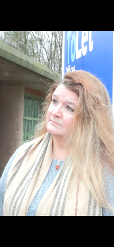 Cheryl Blake Woman with long hair, wearing a scarf, standing near a toilet sign outdoors.