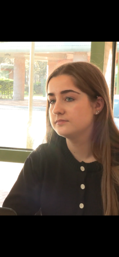 Carrie Baker A young woman in a black top, looking thoughtfully out of a window.