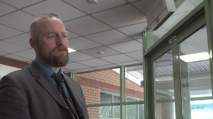 Ethan Fletcher Man in a suit stands in an office corridor, looking thoughtfully towards the window.
