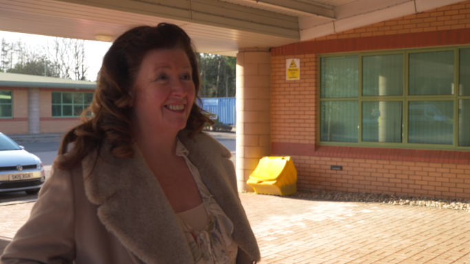 Angie Grant Smiling woman in a light coat stands outside a building with a green window.