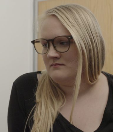 Nicola Thomson Young woman with long blonde hair and glasses, looking thoughtfully to the side.