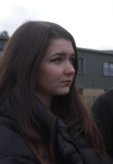 Debbie Grant A young woman with long dark hair, wearing a black jacket, appears contemplative outdoors.